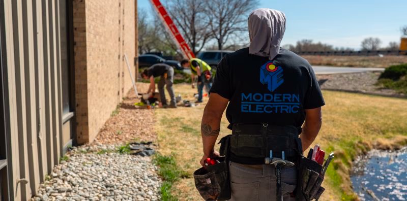 Modern Electric electrician in a branded shirt and tool belt at a Colorado commercial job site with crew working nearby